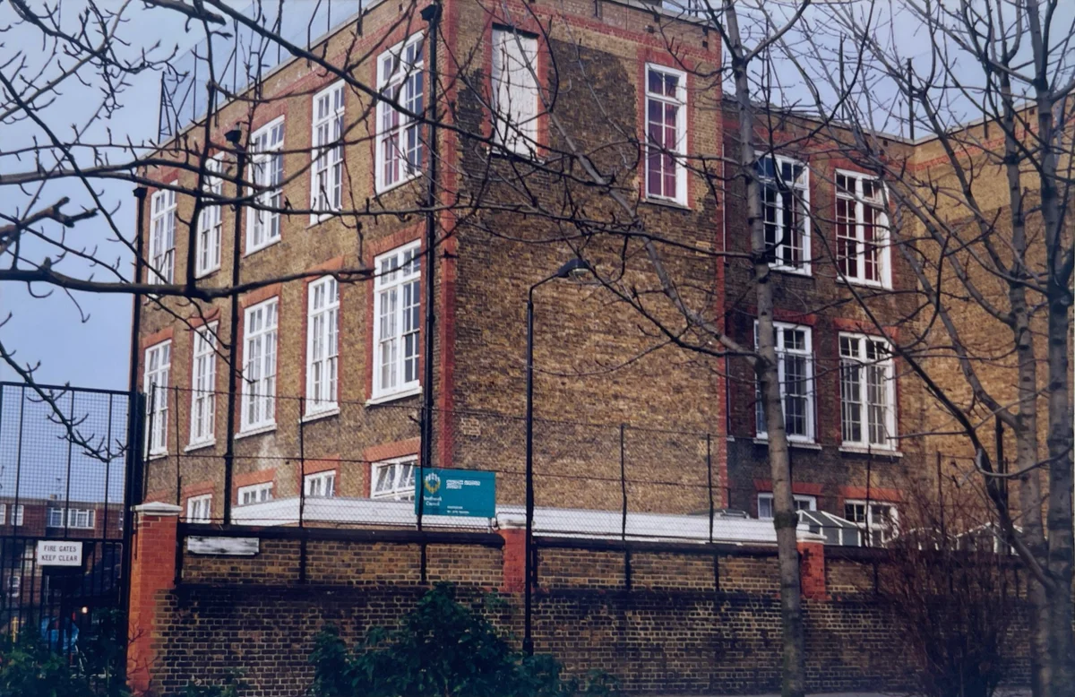 The exterior of a multi-storey Victorian brick school building framed by bare tree branches behind a perimeter fence.