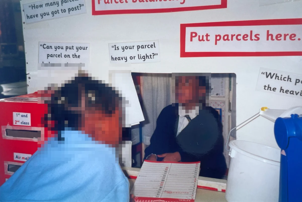 Two young pupils engaged in role-play at a classroom post office counter with educational speech prompts and forms to write on..