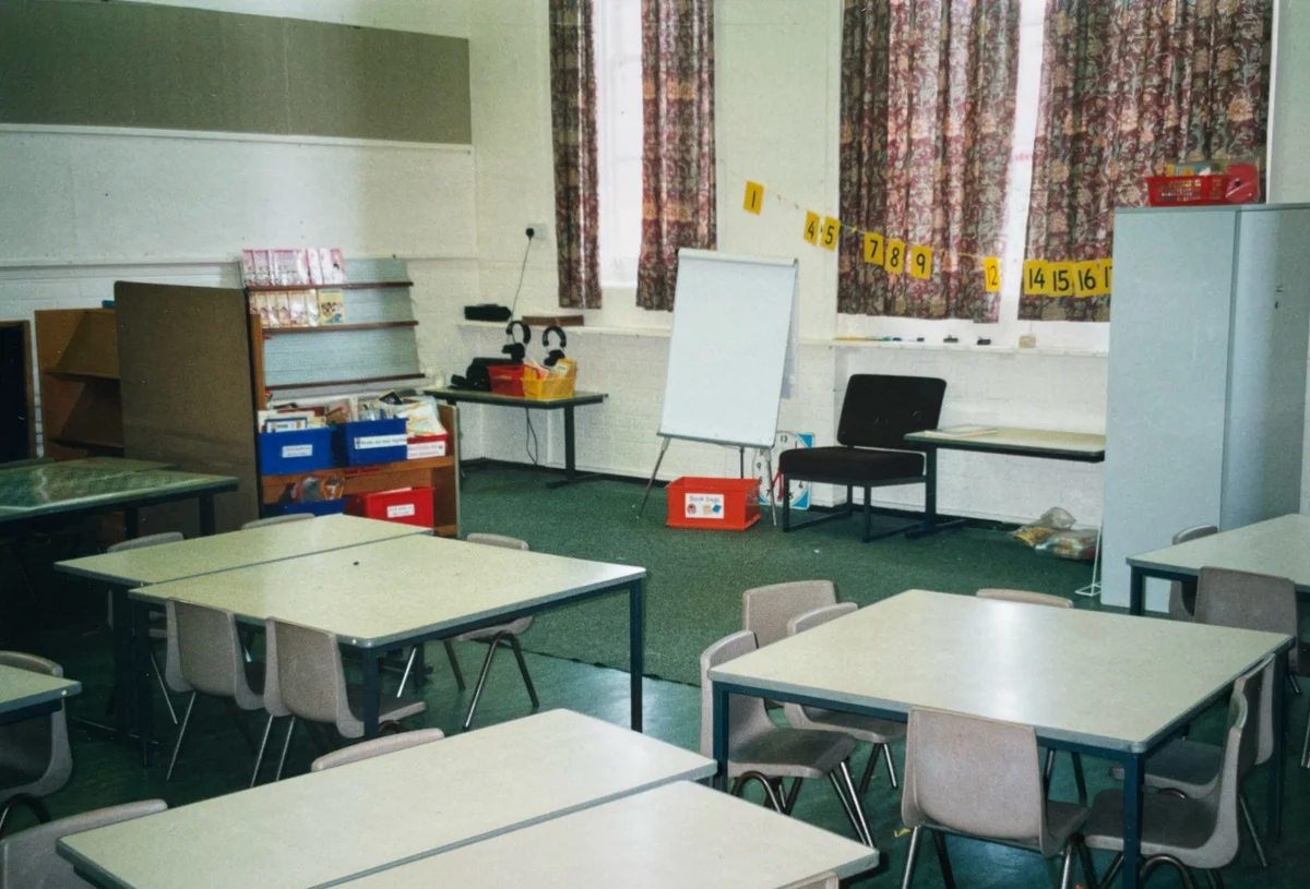 A plain, empty primary school classroom filled with grey and brown furniture, muted flooring, and blank display spaces.