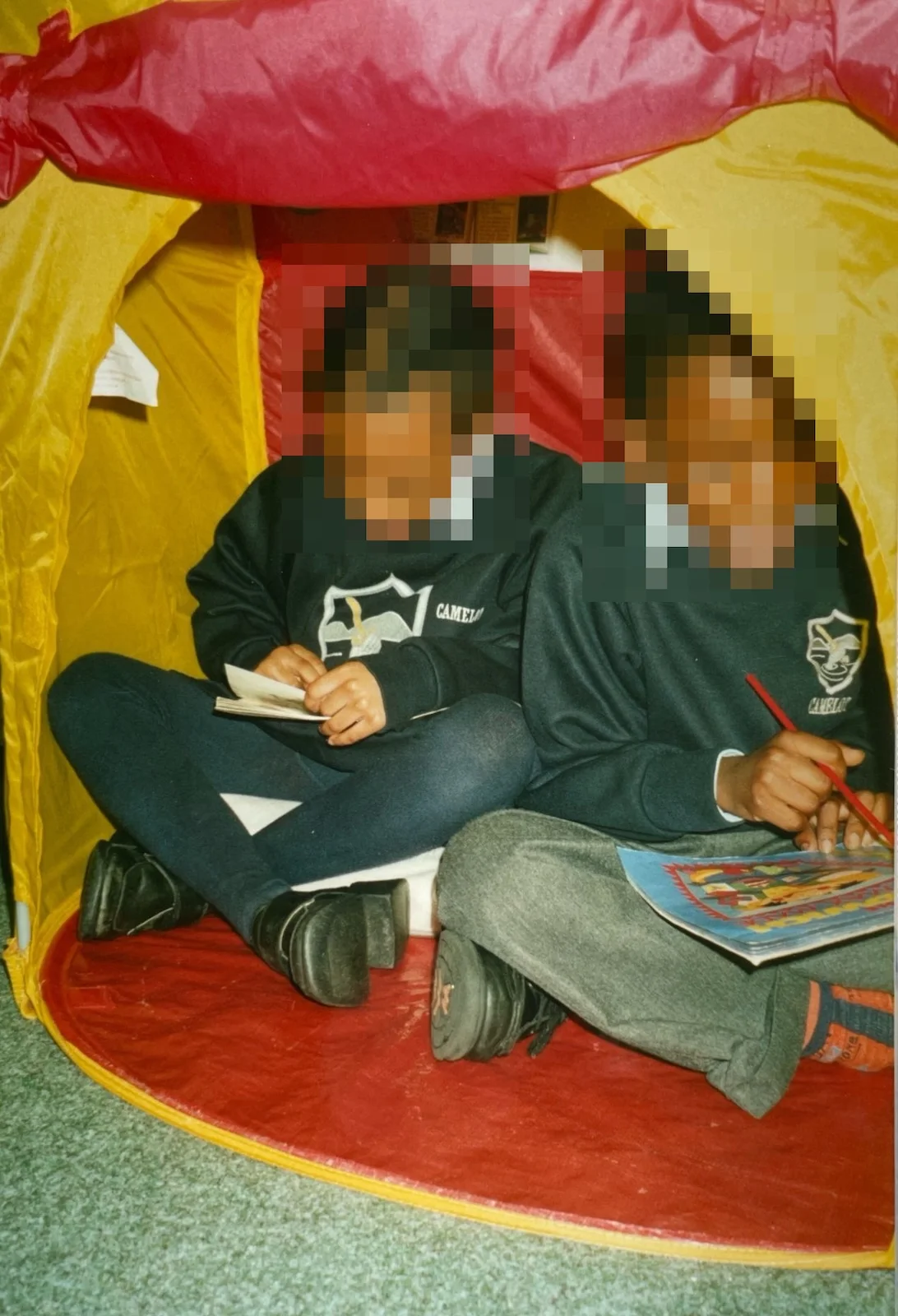 Two young pupils in school uniform sitting cross-legged inside a yellow play tent, one reading a book and the other holding a reading pointer.