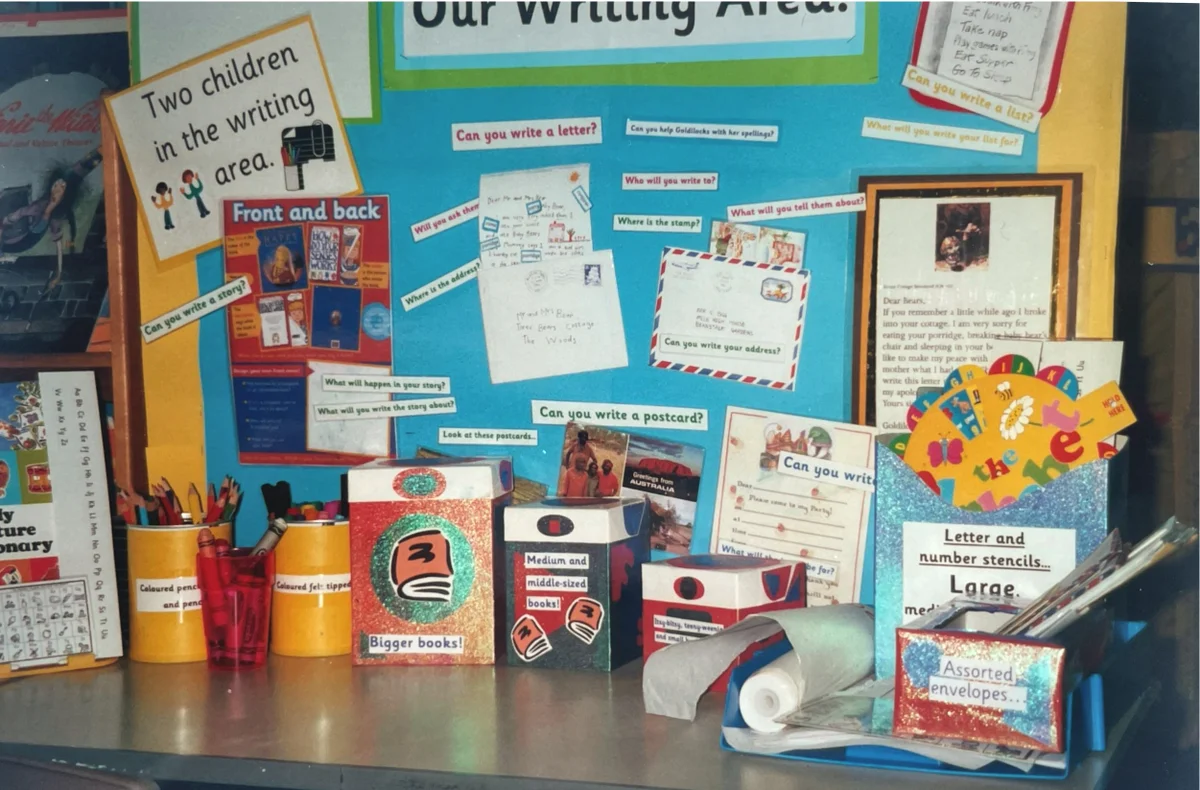 A colourful early years classroom writing area with labelled pots of pencils, stencils, letter paper, and a display board.