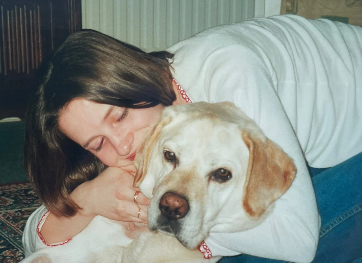 A young woman lying down and warmly hugging a yellow Labrador Retriever indoors.