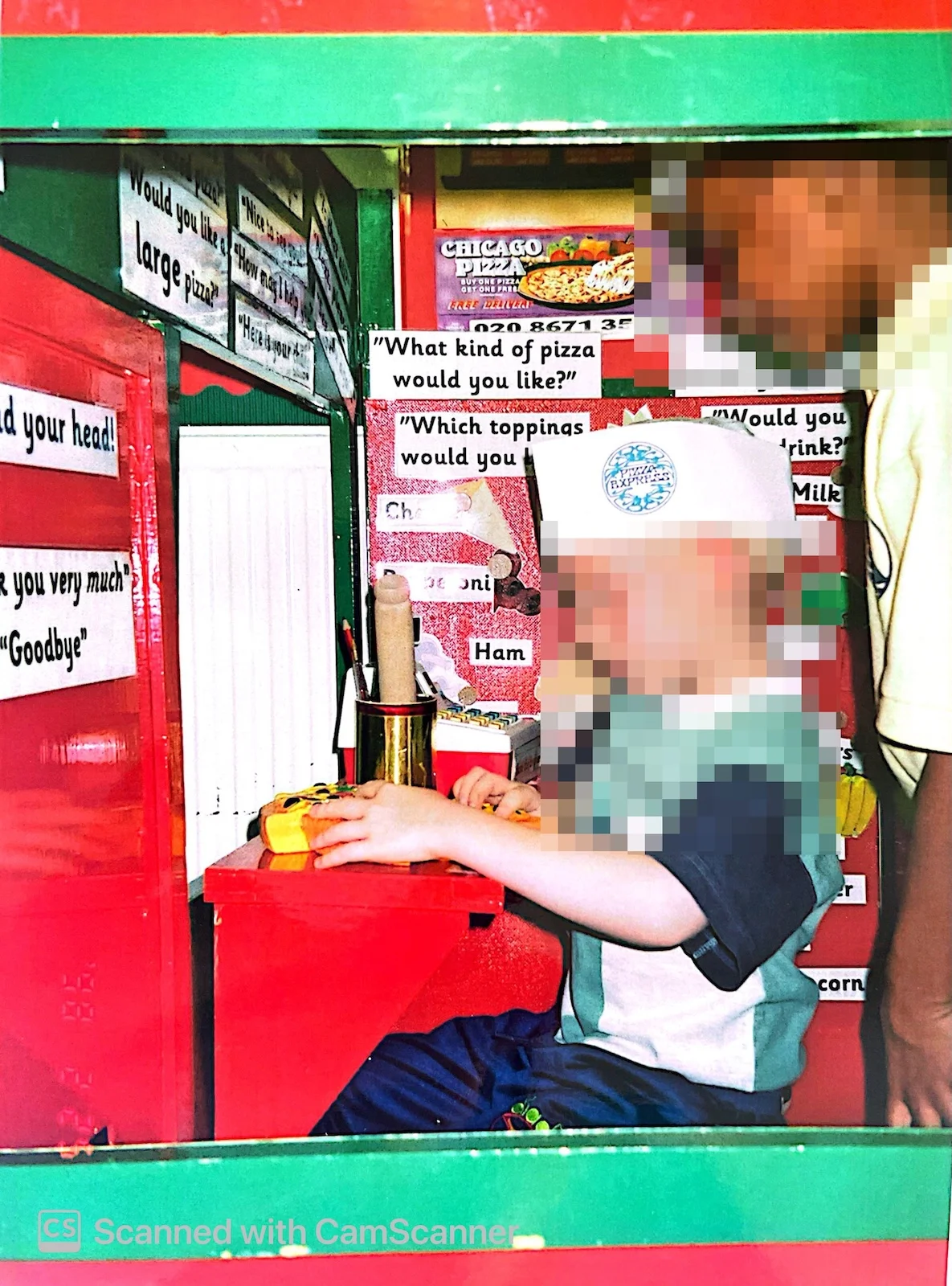 A young child wearing a paper chef hat taking orders in a classroom pizza restaurant roleplay area.