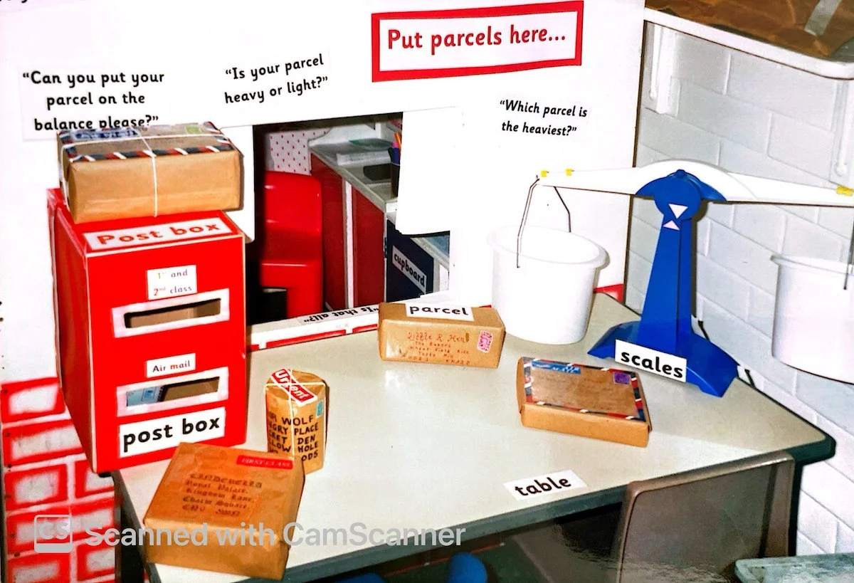 A classroom post office weighing area featuring balance scales, parcel props, a red postbox, and printed prompts.