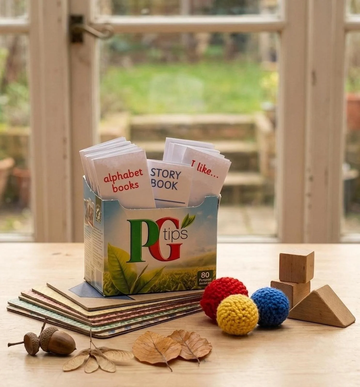 Upcycled PG Tips tea box filled with handmade mini-books alongside acorns, autumn leaves, handmade pompoms, and wooden blocks.