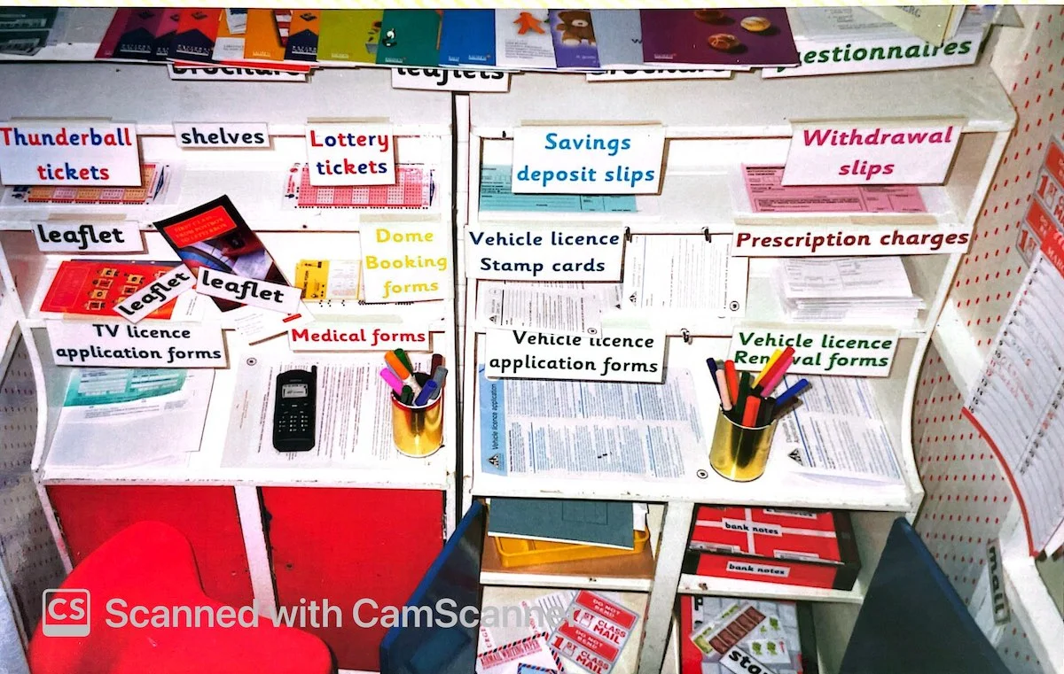 A classroom roleplay post office counter filled with clearly labelled shelves, forms, leaflets, and writing stationery.