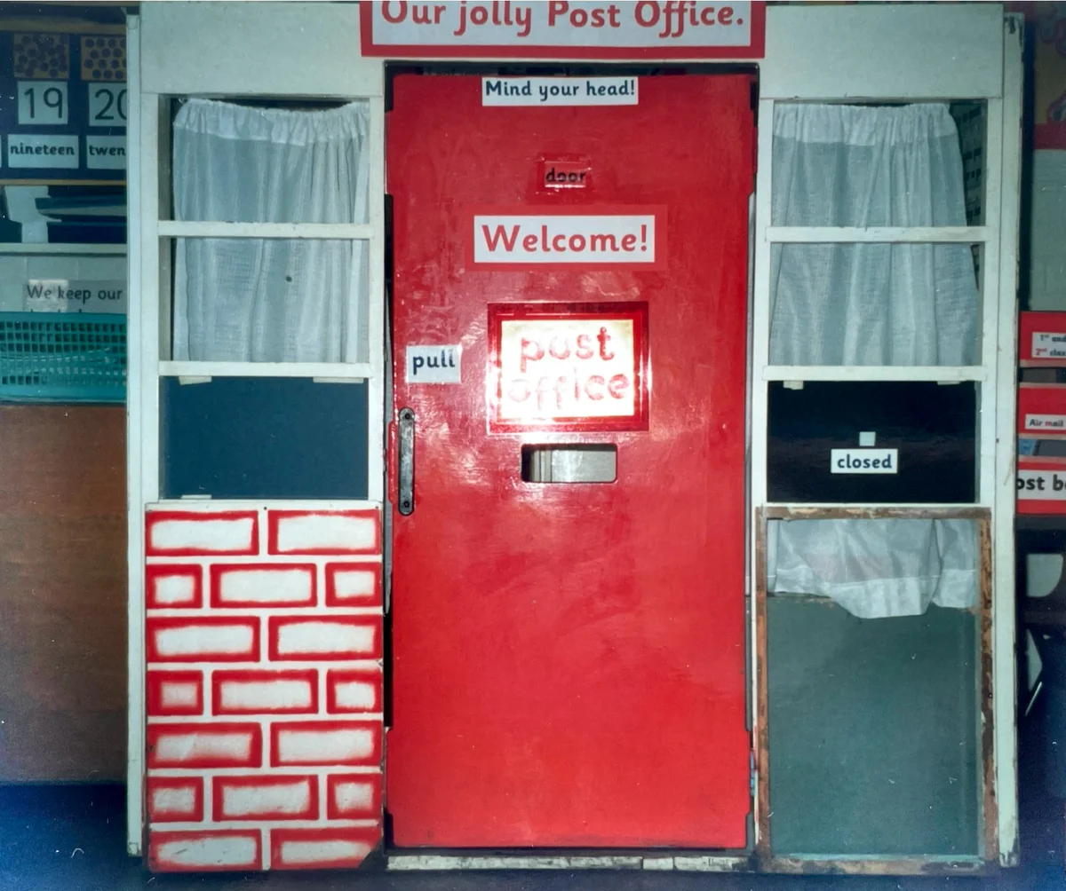 A classroom role-play post office storefront with a red door, white curtains, and brick-patterned panels.