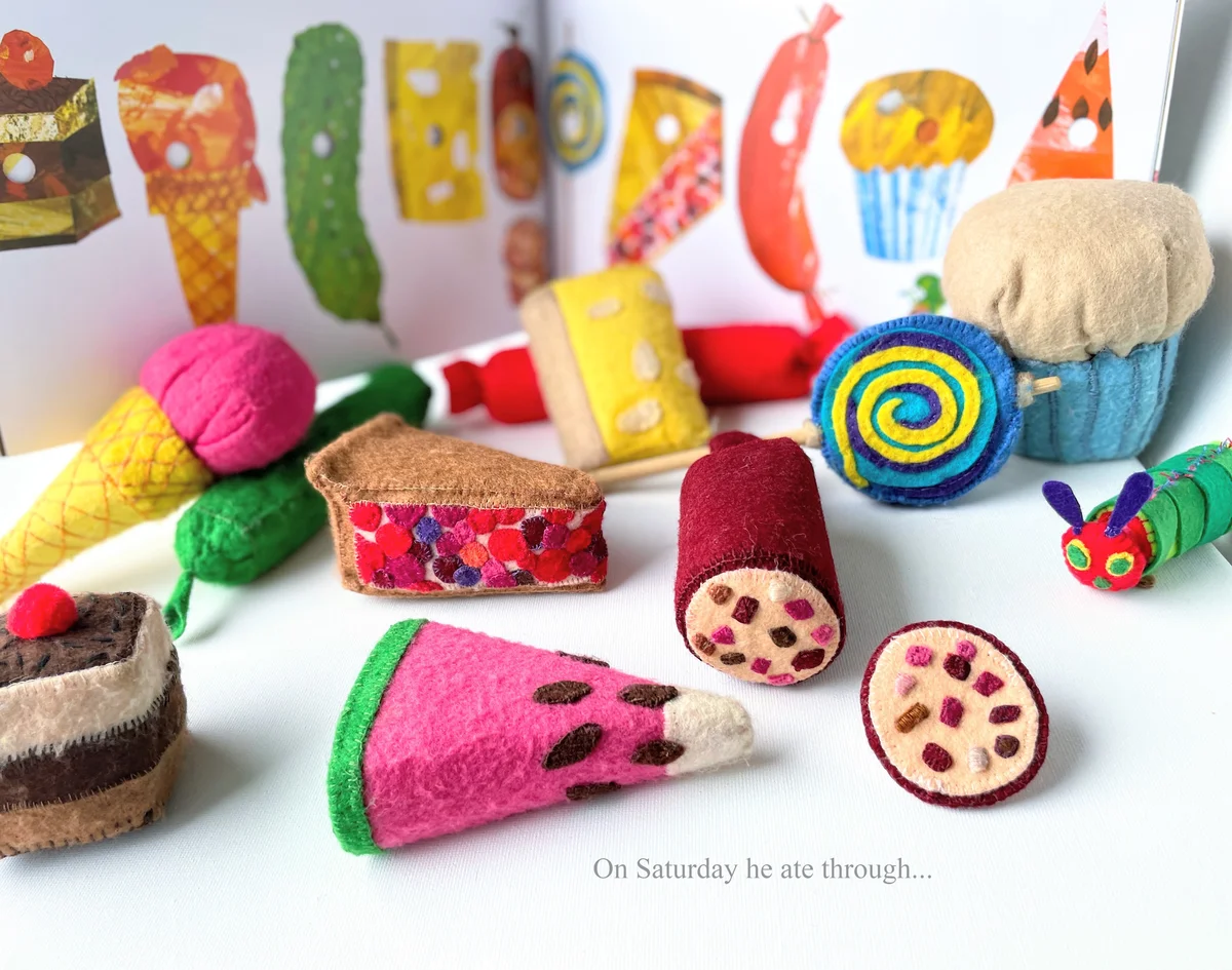 Handmade felt play food items representing the Saturday feast from The Very Hungry Caterpillar, alongside the book and a small caterpillar toy.